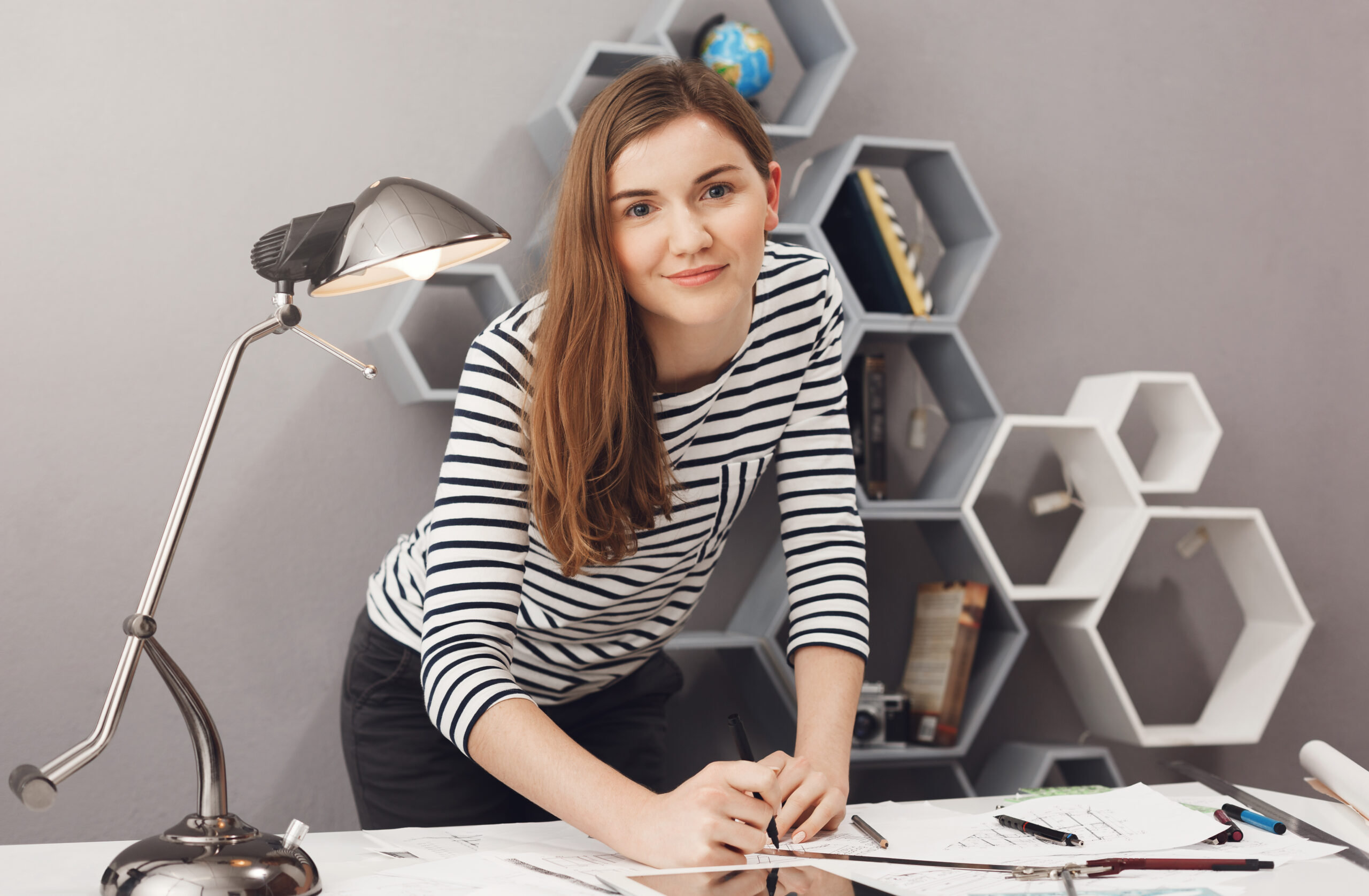 Close up portrait of young charming joyful engineer student girl standing near table, holding hands on work papers, looking in camera with happy and satisfied expression.